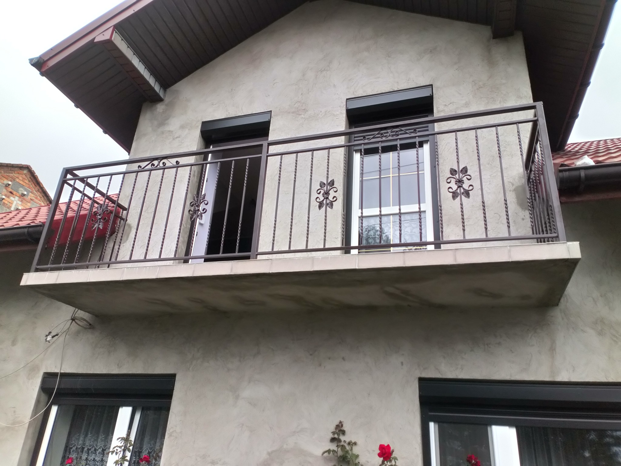 Balkon z metalową balustradą o dekoracyjnych elementach kwiatowych, widok z dołu na elewację budynku z oknami i roletami zewnętrznymi.