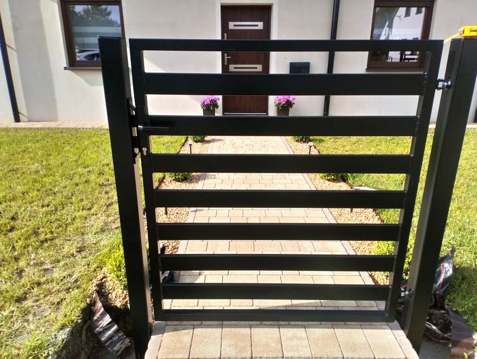 Nowoczesna, czarna furtka z poziomych profili zamontowana w Wieruszowie, widok na ścieżkę z kostki prowadzącą do drzwi wejściowych domu.