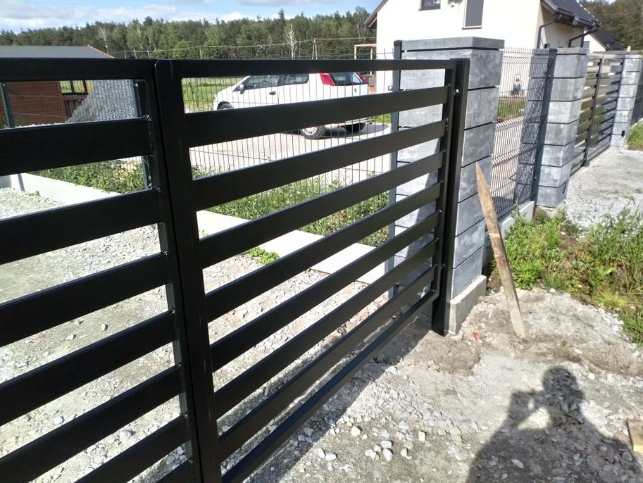 Nowoczesna, czarna brama wjazdowa z poziomymi profilami, zamontowana na betonowych słupkach z szarej kostki, widok z zewnątrz posesji, w tle dom jednorodzinny.