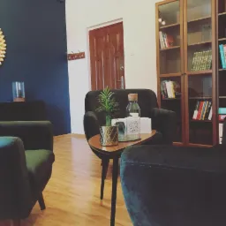 Przytulny gabinet z ciemnozielonymi fotelami, drewnianym regałem z książkami i dekoracyjnym lustrem słonecznym na ciemnoniebieskiej ścianie.