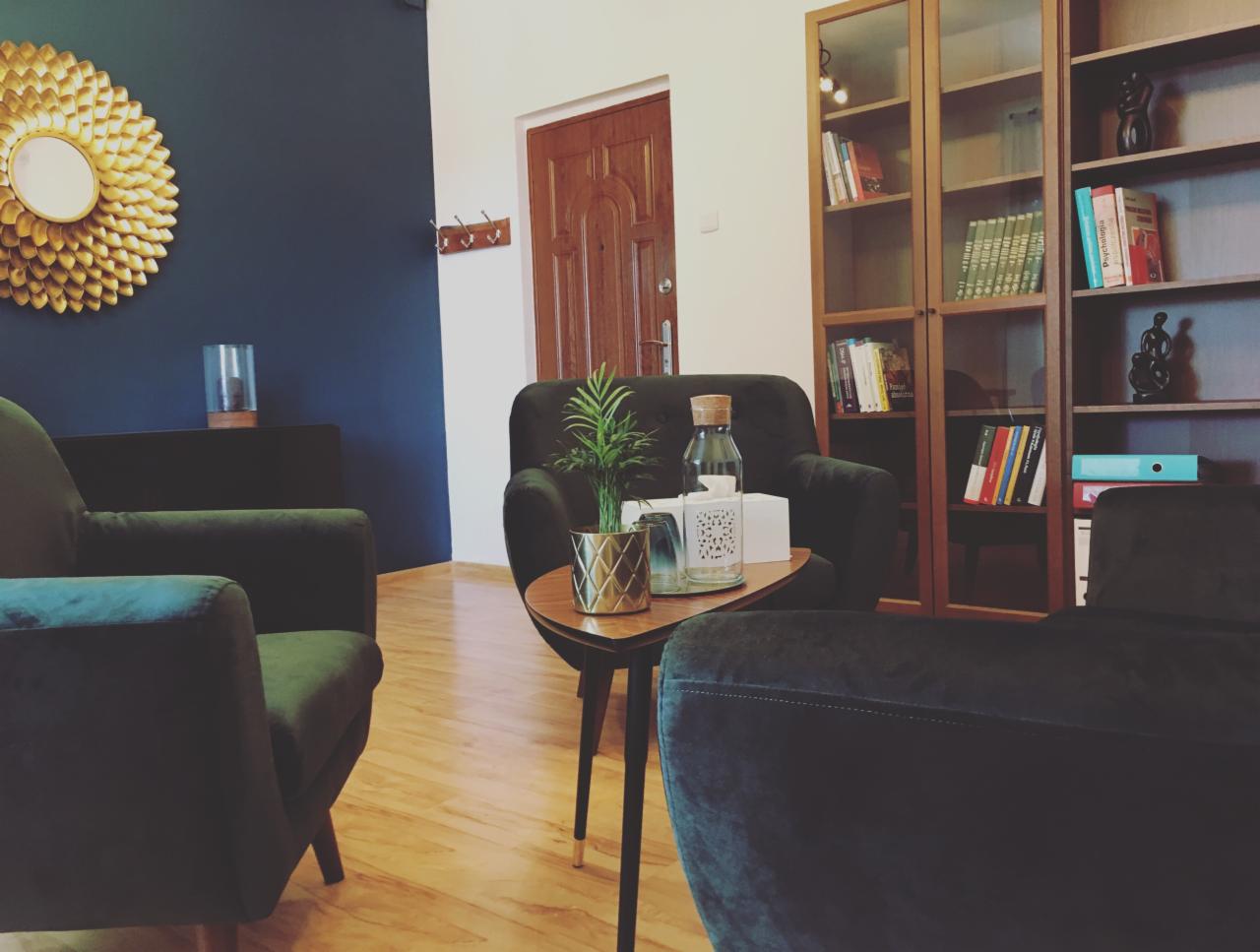 Przytulny gabinet z ciemnozielonymi fotelami, drewnianym regałem z książkami i dekoracyjnym lustrem słonecznym na ciemnoniebieskiej ścianie.