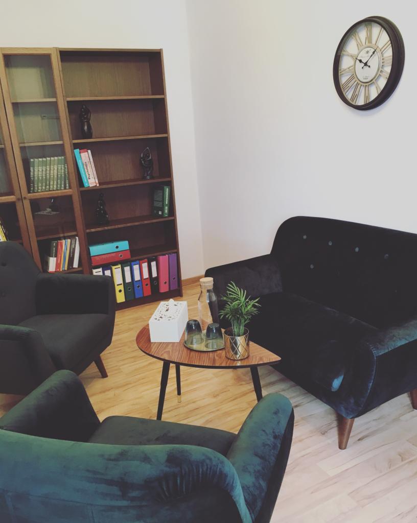 Kącik terapeutyczny: ciemnozielone fotele i sofa, stolik z wodą, szklankami i rośliną, regał z książkami i zegar na ścianie tworzą przytulną przestrzeń.