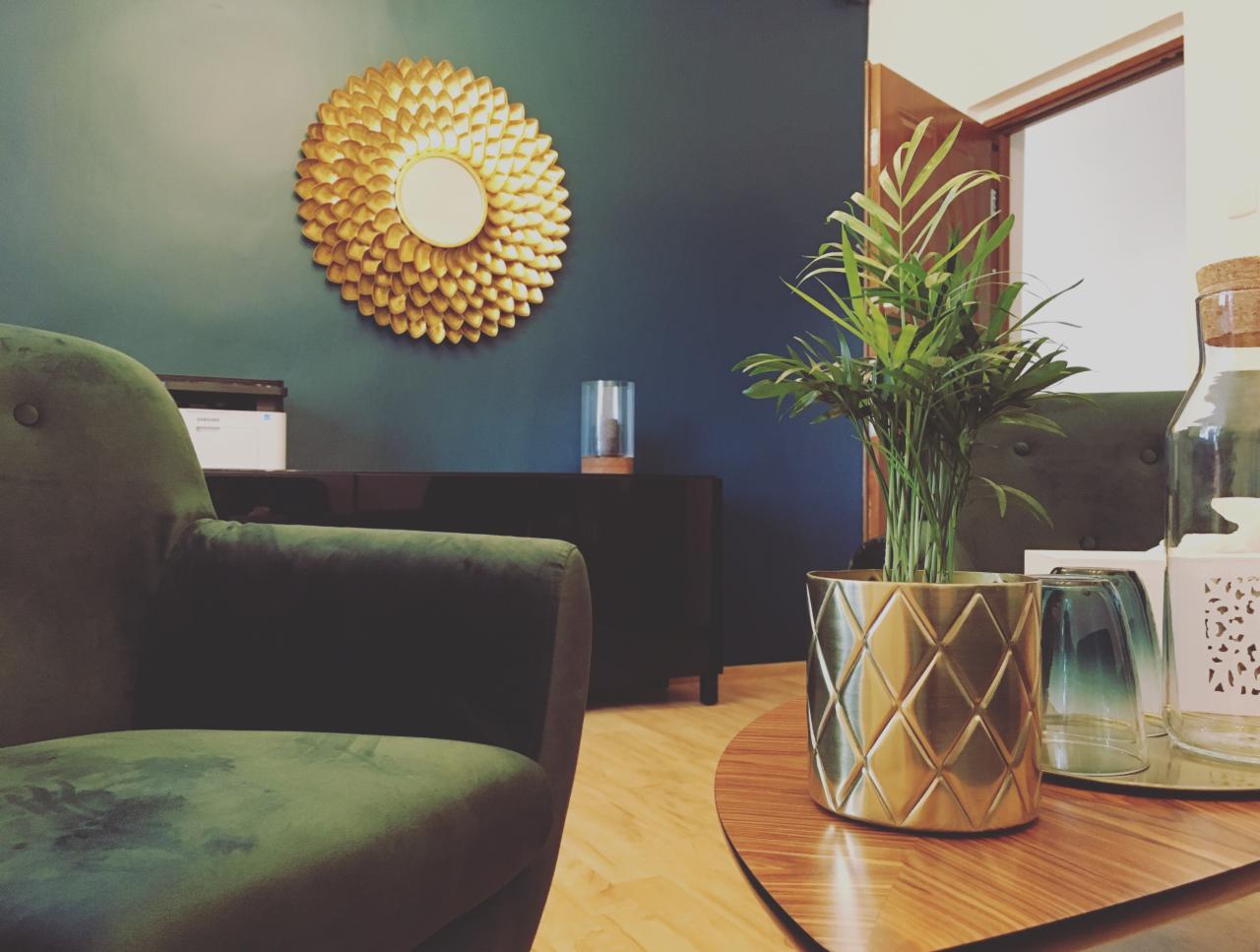 Pokój poczekalni z zielonym welurowym fotelem, okrągłym lustrem w złotej ramie i rośliną w złotej doniczce na okrągłym stoliku kawowym.