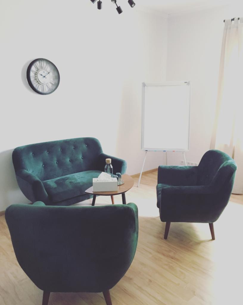 Pokój z zielonymi, welurowymi fotelami i kanapą, stolikiem z wodą i chusteczkami, zegarem na ścianie i tablicą suchościeralną w tle.