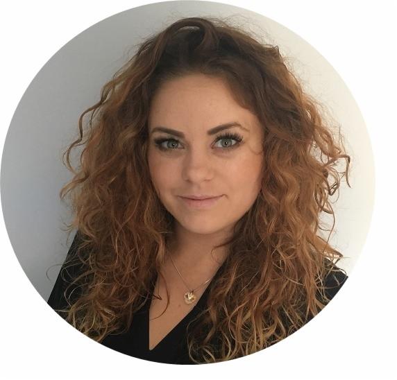 Portret kobiety z kręconymi, rudymi włosami, delikatnym makijażem i złotym naszyjnikiem na tle jasnej ściany.