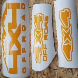 Trzy arkusze białej folii z wyciętymi pomarańczowymi naklejkami z napisem 'LXT OFF ROAD' i grafiką symbolizującą pojazd terenowy, ułożone na drewnianej powierzchni.