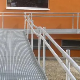 Zewnętrzna rampa dla wózków inwalidzkich wykonana z kraty pomostowej, z metalowymi poręczami, prowadząca do budynku o pomarańczowej elewacji.