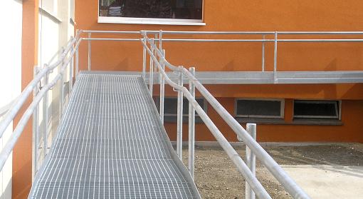 Zewnętrzna rampa dla wózków inwalidzkich wykonana z kraty pomostowej, z metalowymi poręczami, prowadząca do budynku o pomarańczowej elewacji.