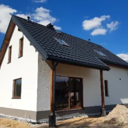 Nowoczesny dom jednorodzinny z ciemnoszarą dachówką, białymi ścianami i drewnianymi elementami wykończeniowymi, widoczny w słoneczny dzień na tle błękitnego nieba z chmurami.