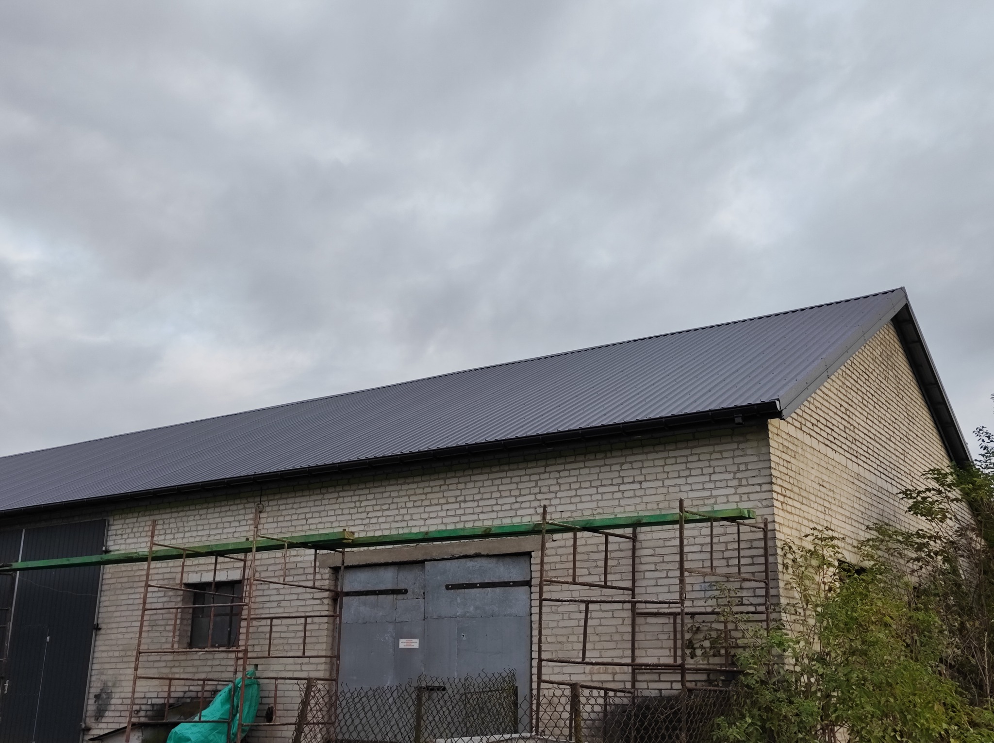 Budynek gospodarczy z jasnej cegły z nowym, ciemnoszarym dachem z blachy trapezowej, częściowo zasłonięty rusztowaniami i zieloną plandeką, pochmurne niebo w tle.