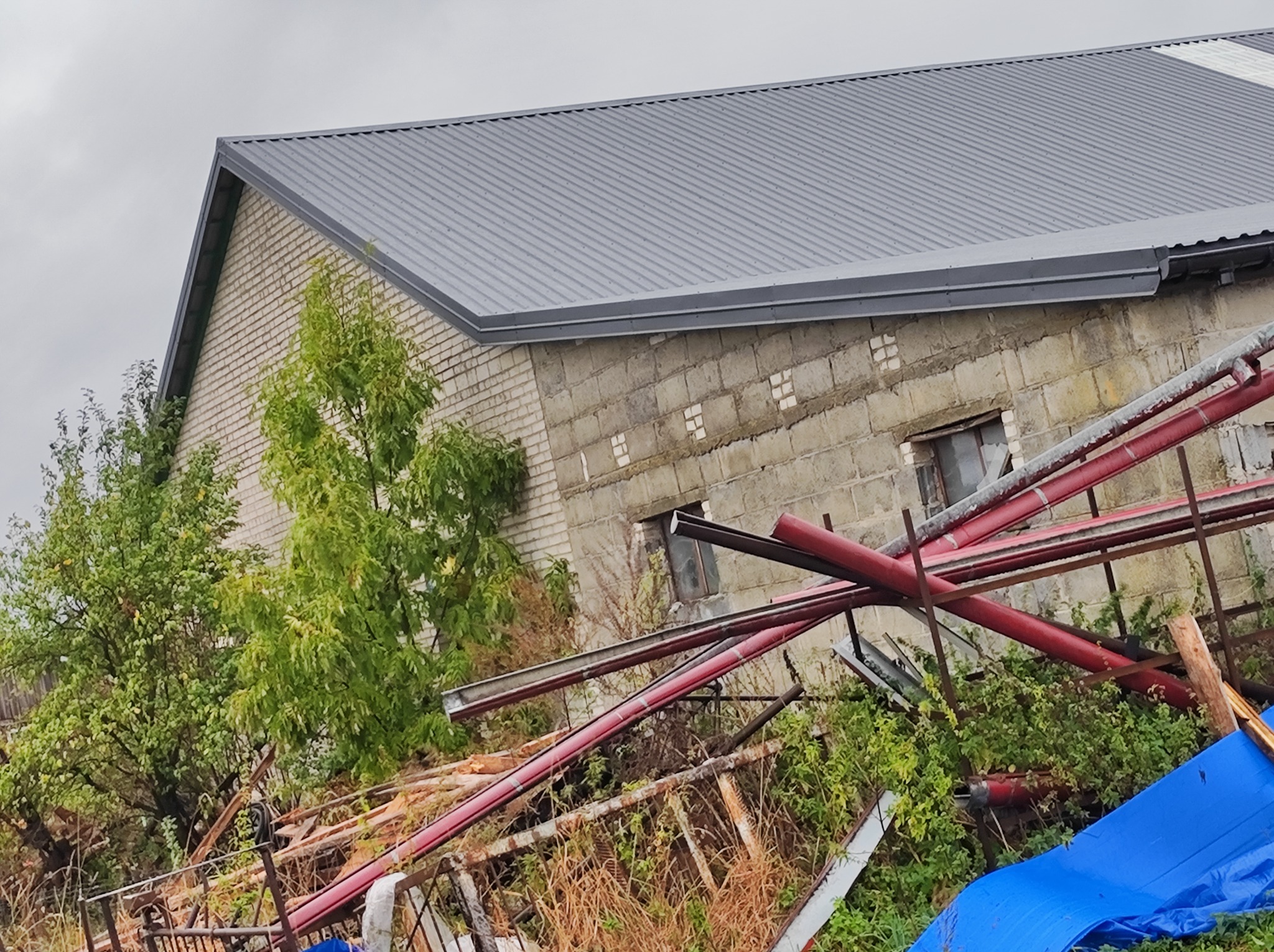 Szary dach z blachy trapezowej na jednopiętrowym budynku z białej cegły, widoczny fragment zniszczonej posesji z rurami i niebieską plandeką w nieładzie.