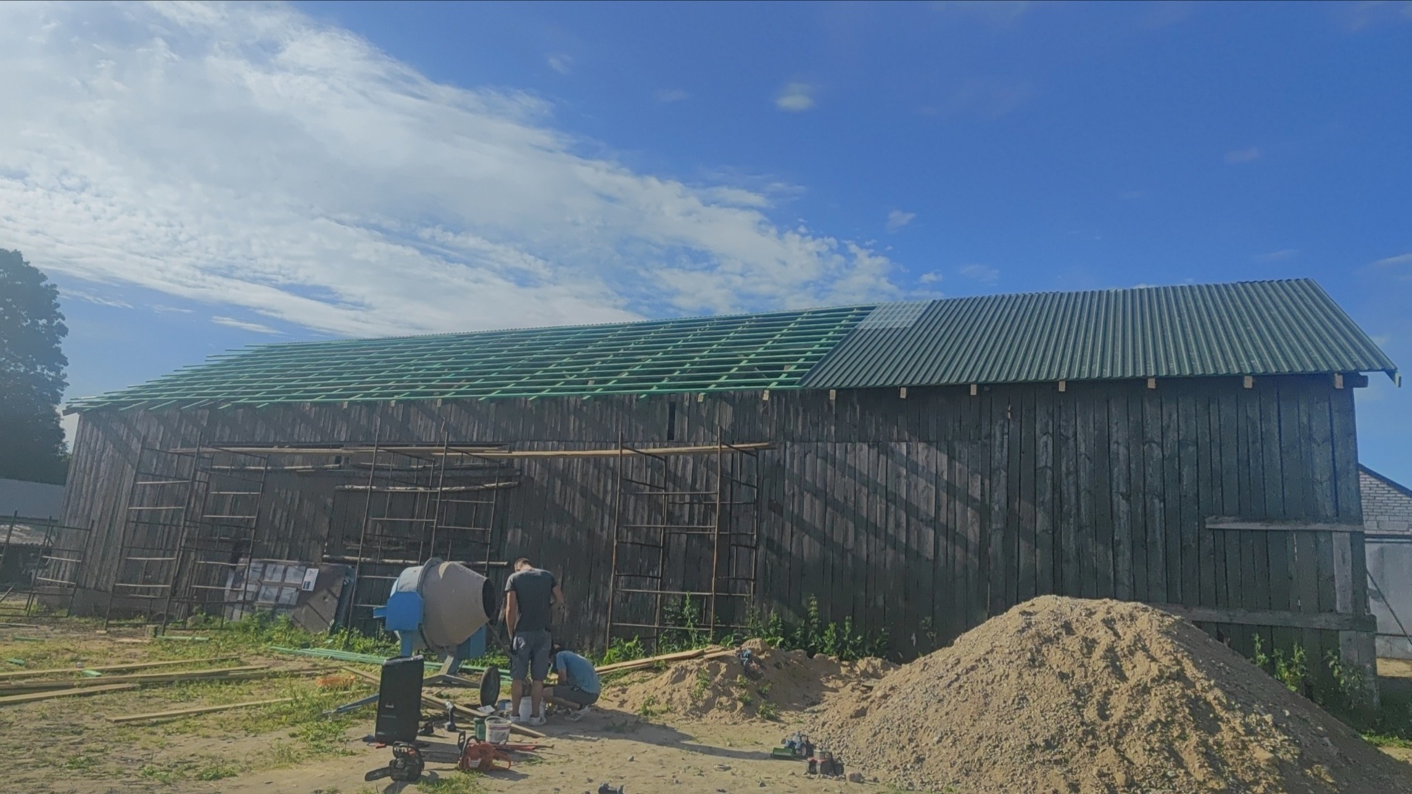 Remont dachu starego, drewnianego budynku gospodarczego z częściowo ułożoną konstrukcją i blachą trapezową, widoczna betoniarka i ekipa budowlana w trakcie prac.