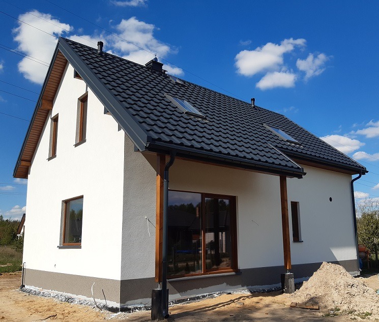 Nowoczesny dom jednorodzinny z ciemnoszarą dachówką, białymi ścianami i drewnianymi elementami wykończeniowymi, widoczny w słoneczny dzień na tle błękitnego nieba z chmurami.