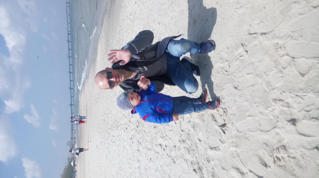 Mężczyzna w okularach przeciwsłonecznych leży na piasku z małym dzieckiem w niebieskiej kurtce, w tle widok na morze i molo.