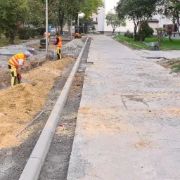 Prace ziemne w Białobrzegach: robotnicy w odblaskowych kamizelkach przygotowują teren pod nową nawierzchnię chodnika, widoczny wykop, obrzeża i ciężki sprzęt budowlany.