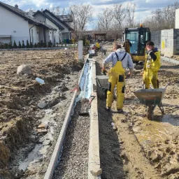 Budowa obrzeża z kostki betonowej z drenażem żwirowym, dwóch pracowników w żółtych kombinezonach oraz mini koparka w tle, teren budowy w błocie.