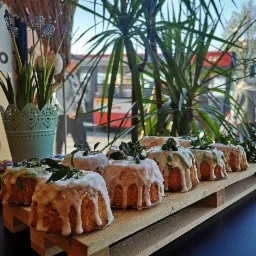 Rząd małych babek wielkanocnych z białym lukrem i zielonymi listkami na drewnianej palecie, w tle roślina doniczkowa i widok na ulicę przez szybę.