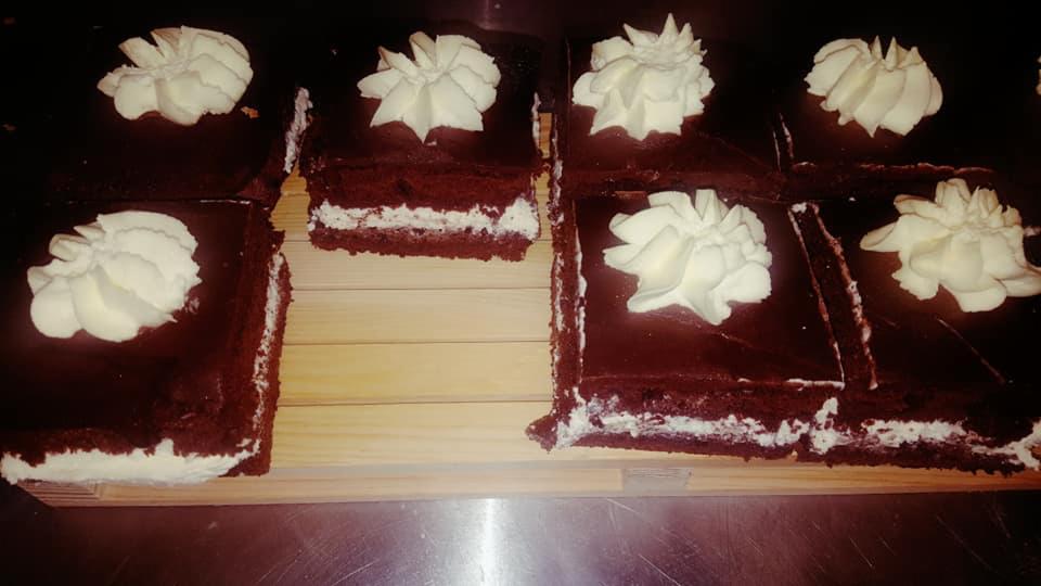 Kawałki czekoladowego ciasta z kremem i polewą, udekorowane rozetkami z bitej śmietany, ułożone na drewnianej desce.