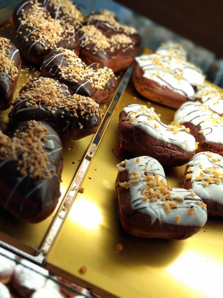 Słodkie pączki w kształcie serca, część oblana ciemną czekoladą i posypana orzechami, część białą polewą z ciemnymi paskami i posypką, ułożone na złotej tacy.