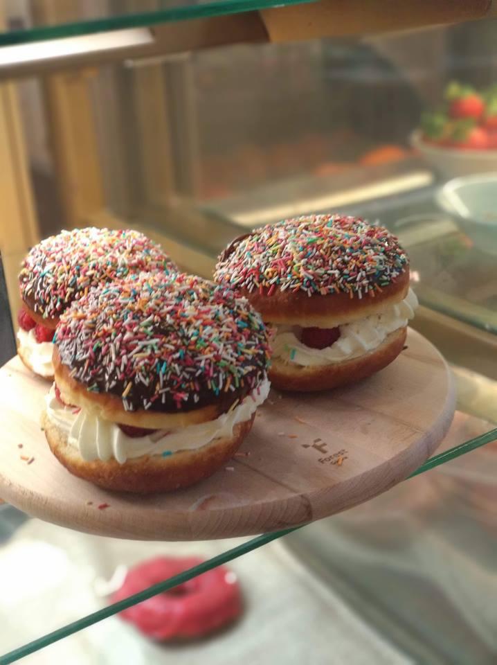 Trzy pączki w stylu amerykańskim, z kremem, malinami i polewą czekoladową posypaną kolorową posypką, ułożone na drewnianej tacy w witrynie cukierniczej.