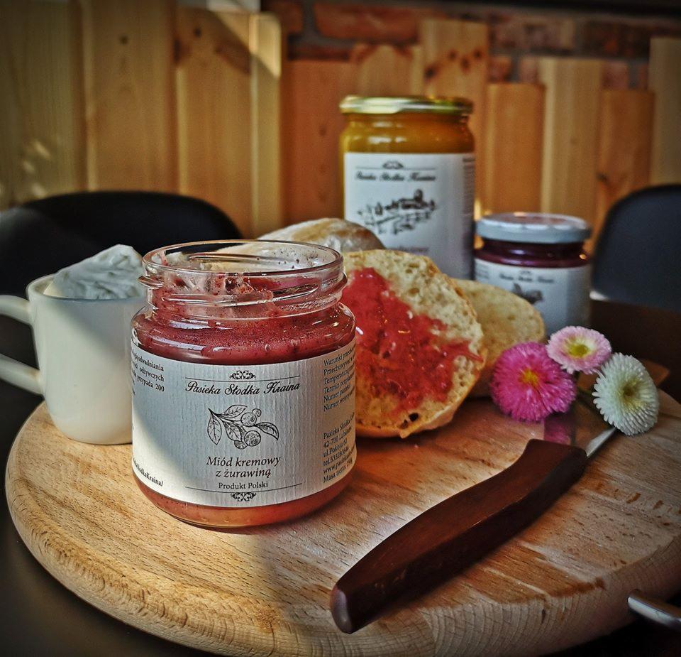 Słoik kremowego miodu żurawinowego marki Pasieka Słodka Kraina na drewnianej desce do krojenia, obok kromki chleba posmarowanej miodem, filiżanki z białym kremem, kwiatów i noża z drewnianą rączką,...