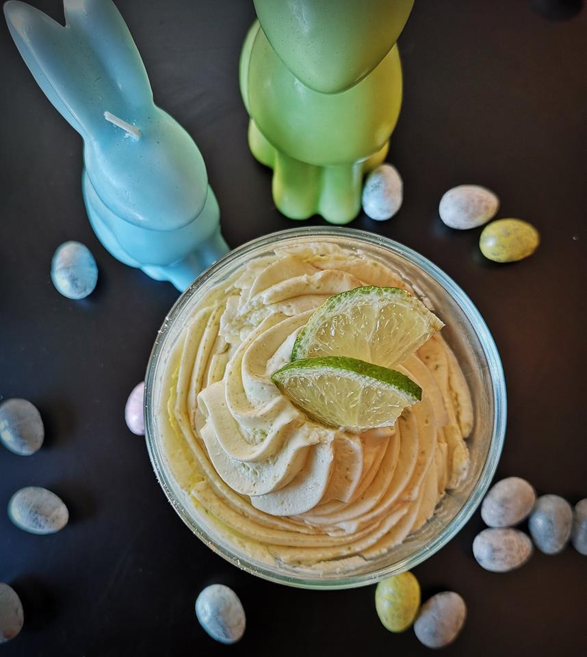 Deser w szklanej miseczce, udekorowany dwoma plasterkami limonki, otoczony pastelowymi jajkami i świeczkami w kształcie zajączków na czarnym tle.