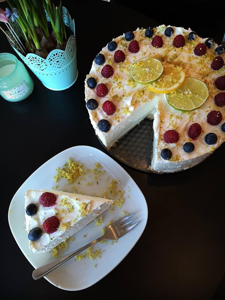 Tort bezowy udekorowany malinami, borówkami i plasterkami limonki, obok wycięty kawałek na talerzu z widelcem, w tle doniczka z kwiatami i świeca.