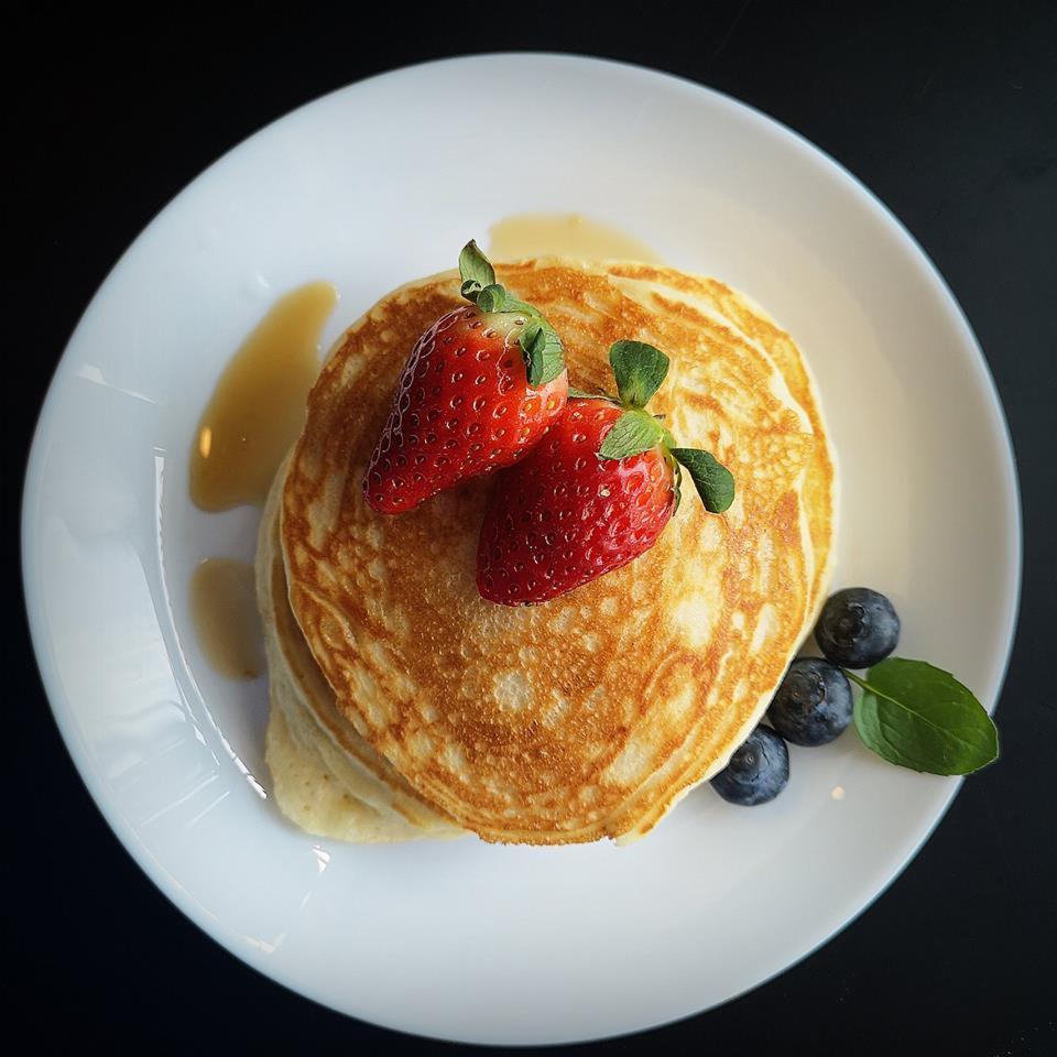 Stos apetycznych, złocistych naleśników na białym talerzu, polanych syropem i udekorowanych świeżymi truskawkami i borówkami, na ciemnym tle.