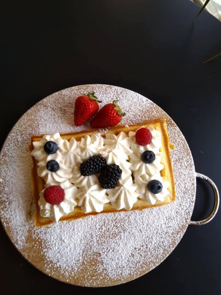 Prostokątny gofr udekorowany bitą śmietaną, borówkami, malinami i jeżynami, posypany cukrem pudrem, ułożony na okrągłej drewnianej desce z uchwytem na czarnym tle.