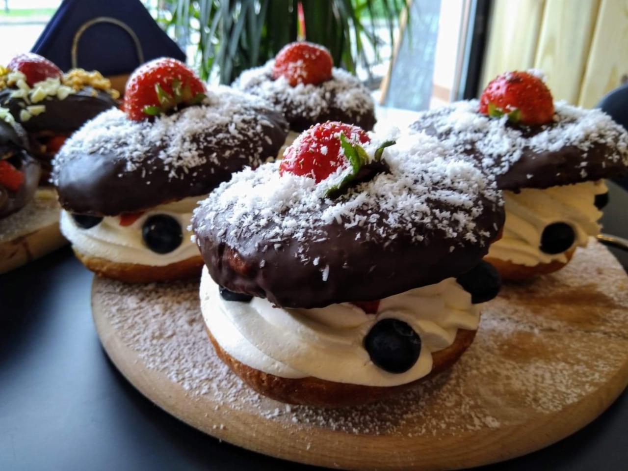 Dekoracyjne pączki z kremem, borówkami, polewą czekoladową i posypką kokosową, zwieńczone świeżymi truskawkami na drewnianej desce.