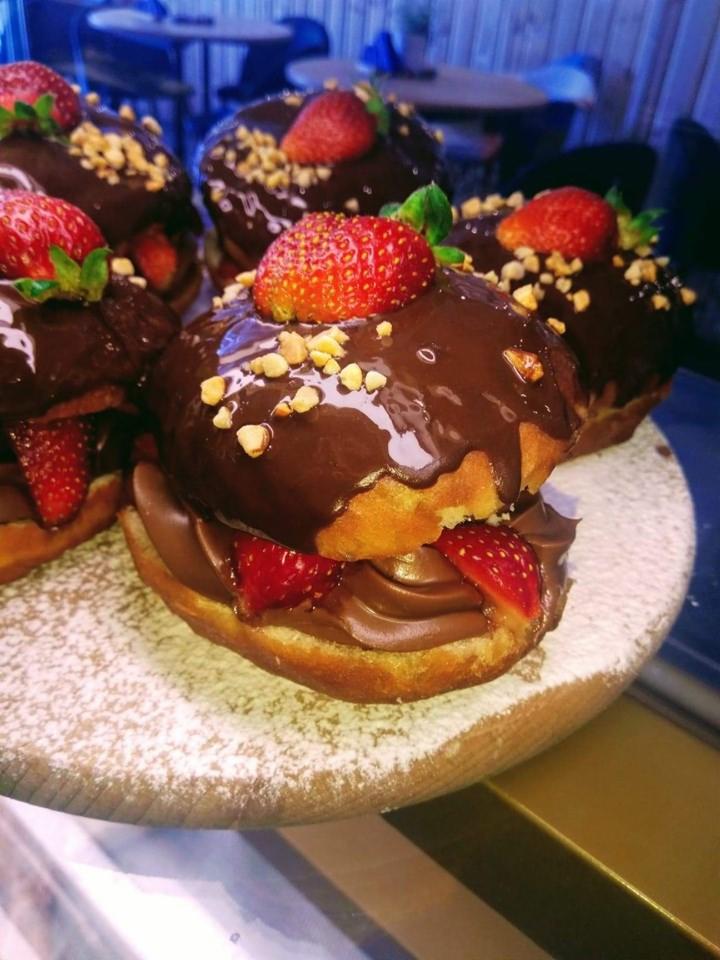 Słodkie donuty z kremem czekoladowym i świeżymi truskawkami, posypane orzechami, ułożone na drewnianej desce oprószonej cukrem pudrem.