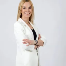 Portret uśmiechniętej kobiety w białym żakiecie, z rękami skrzyżowanymi na piersiach, na jasnym tle.