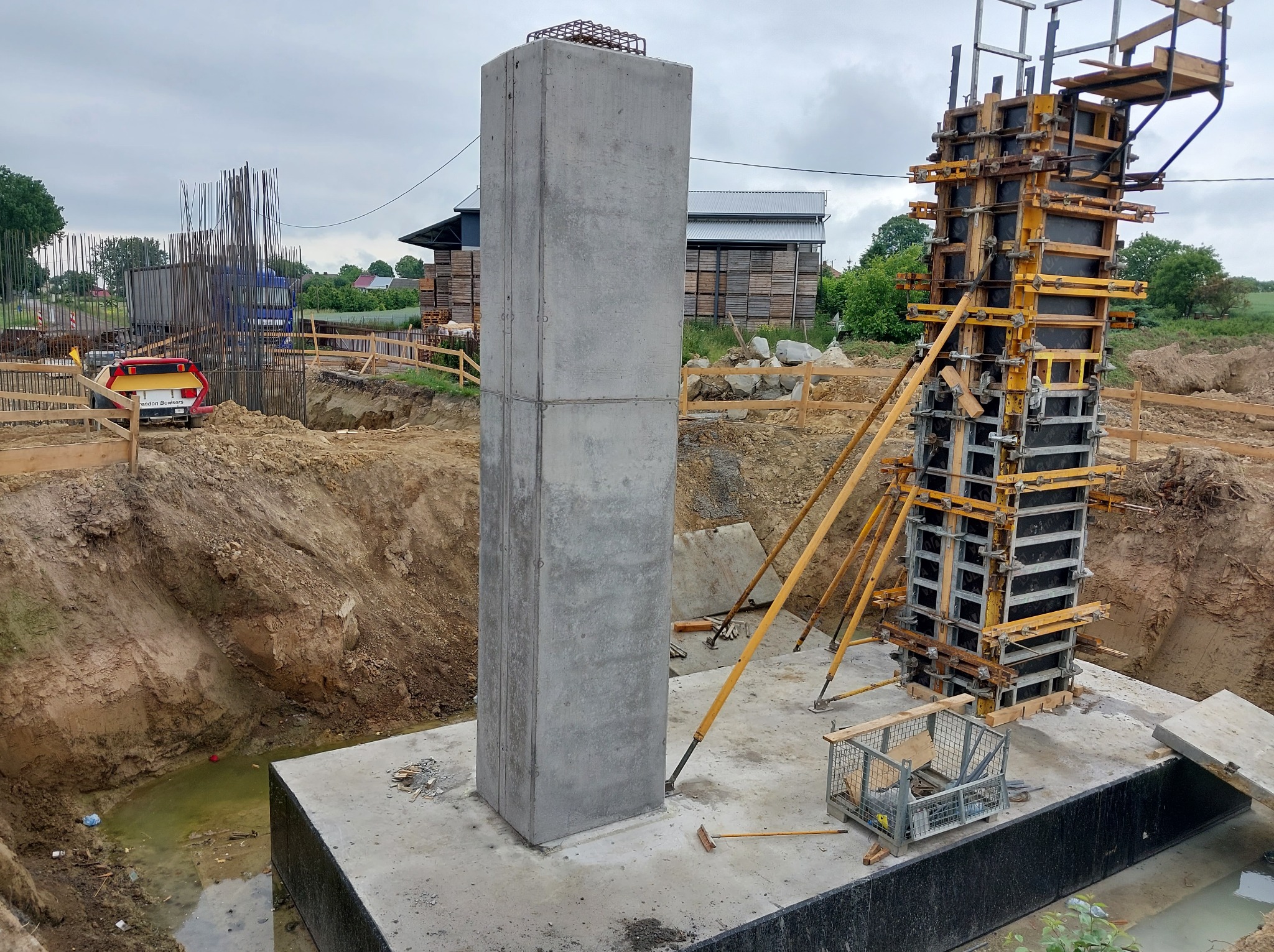 Żelbetowy słup w trakcie budowy, widoczne szalunki i zbrojenie. Słup z betonu, teren budowy z wykopami i sprzętem. Pochmurne niebo w tle.