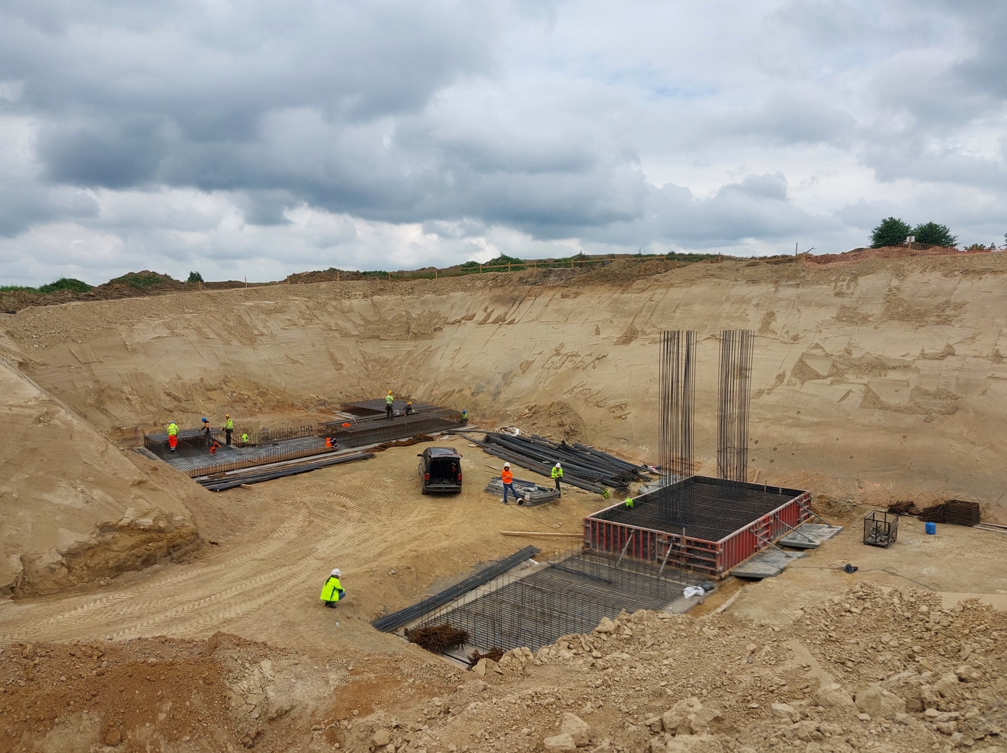 Budowa fundamentów: zbrojenie, szalunki i pracownicy w wykopie. Widok z góry na plac budowy z elementami konstrukcji żelbetowej i pojazdem w tle.