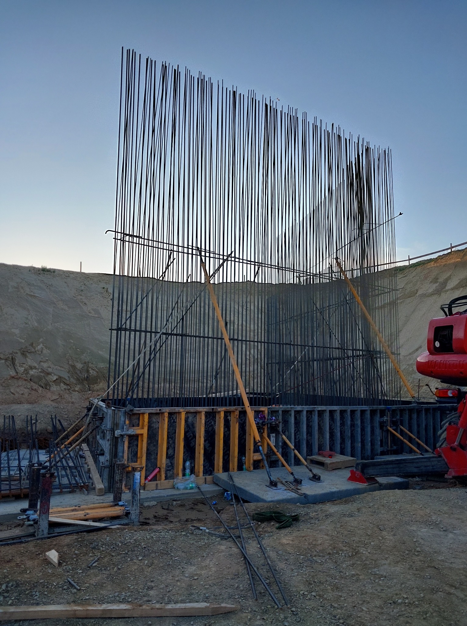 Zbrojenie pionowe w szalunku, wsparte drewnianymi stemplami. Widoczny fragment czerwonej maszyny budowlanej i ziemny nasyp w tle. Ujęcie z dołu, wczesnym rankiem.