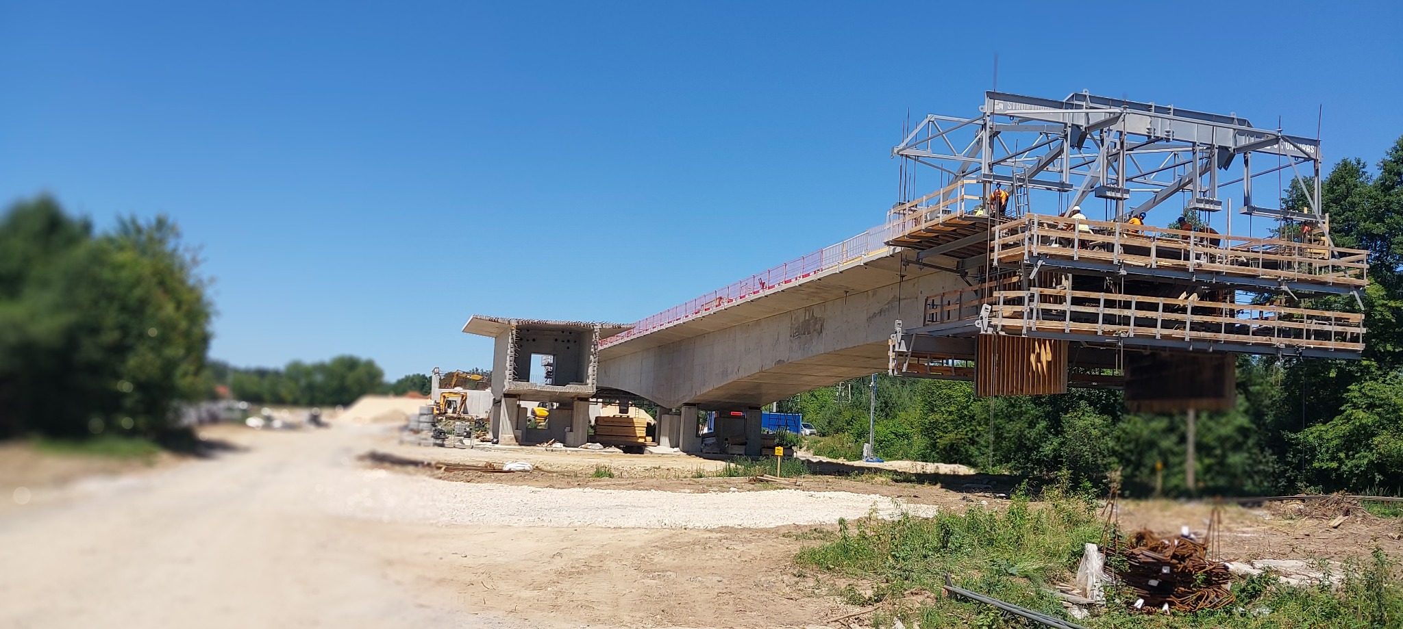 W budowie betonowy wiadukt z widocznym zbrojeniem i pracownikami na rusztowaniu, w tle błękitne niebo i zieleń drzew. Ujęcie w słoneczny dzień.