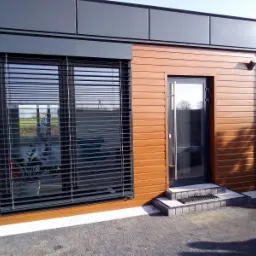 Nowoczesny budynek z brązową elewacją imitującą drewno i szarymi panelami, duże okno z żaluzjami, szklane drzwi wejściowe z trzema kamiennymi stopniami i wycieraczką.