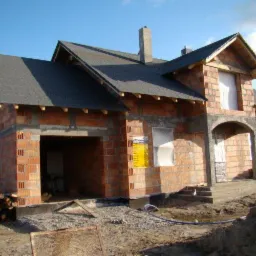 Niedokończony dom jednorodzinny z czerwonej cegły, z ciemnym dachem i widocznym kominem, na etapie budowy, z elementami konstrukcyjnymi w stanie surowym i stertą drewna obok.