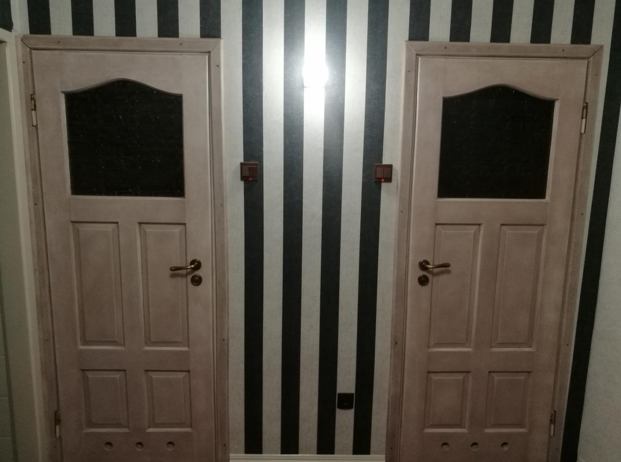 Dwa jasne, drewniane drzwi z szybami, zamontowane na tle ściany w czarno-białe pionowe pasy, z widocznymi włącznikami światła.
