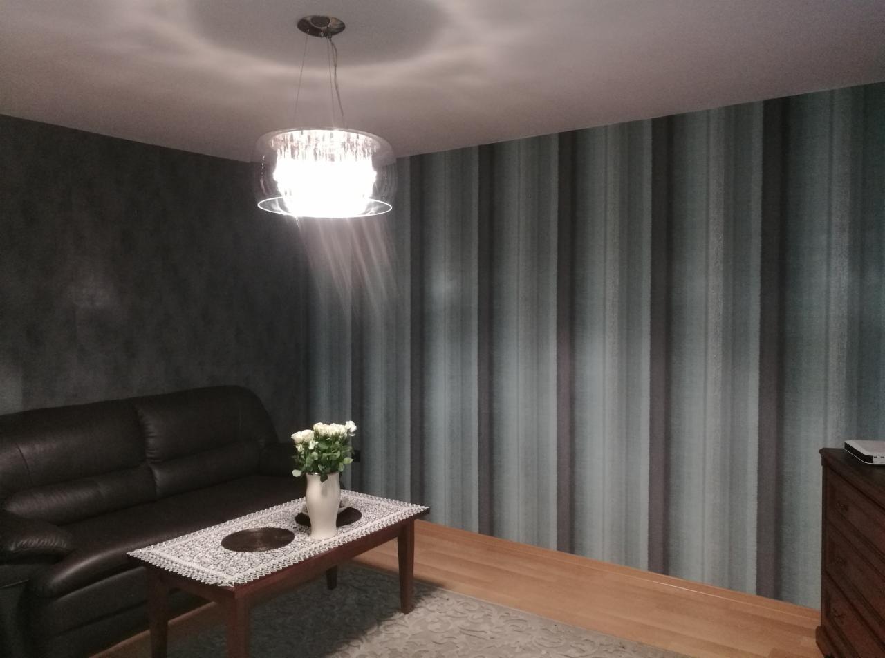 Salon z ciemną skórzaną sofą, stolikiem z kwiatami, lampą sufitową z kryształkami i tapetą w pionowe pasy w odcieniach szarości i błękitu.