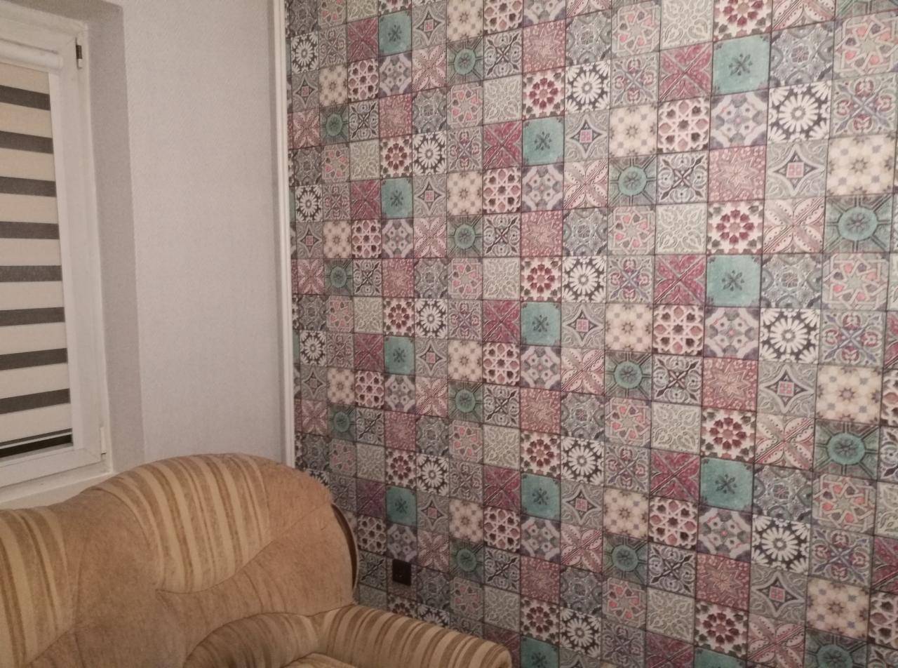 Ściana wykończona wzorzystą tapetą w stylu patchwork, z widocznym fragmentem beżowego fotela w paski i roletami okiennymi w tle.