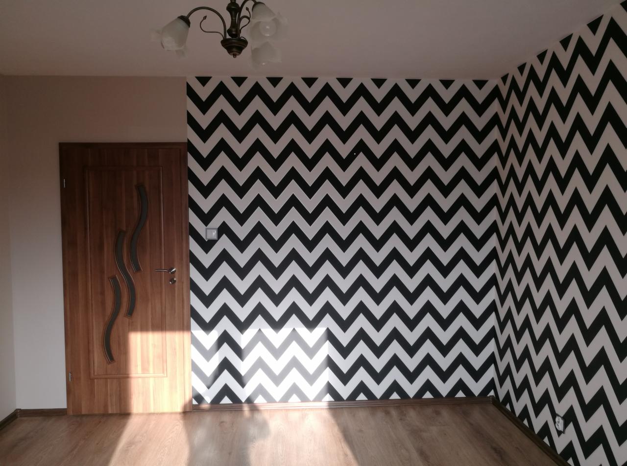 Pokój po odświeżeniu z biało-czarną tapetą w zygzaki na ścianie, widoczna drewniana podłoga i drzwi z dekoracyjnymi wstawkami, oświetlenie sufitowe w stylu retro.