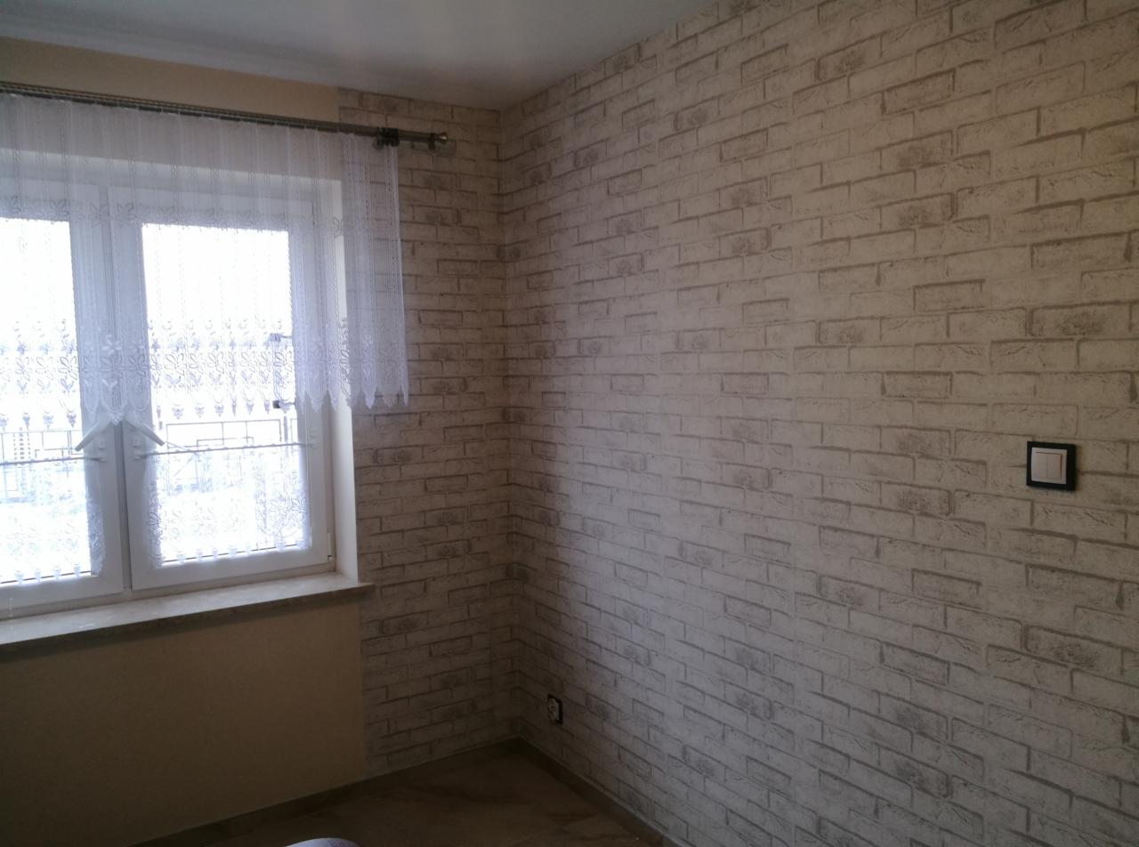 Pokój po odświeżeniu: ściana pokryta tapetą imitującą białą cegłę, okno z białą, koronkową firanką, gniazdko elektryczne na ścianie.