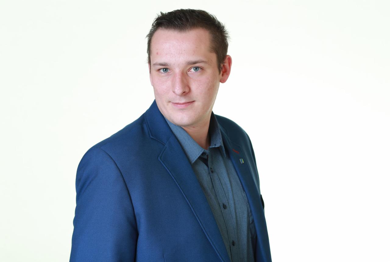 Portret młodego mężczyzny w niebieskiej marynarce i wzorzystej koszuli, uśmiechającego się do kamery na jasnym tle.