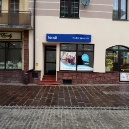 Fasada budynku z szyldem 'lendi Kredyty i pożyczki', obok wejście do sklepu z telefonami komórkowymi, mokry chodnik z kostki brukowej.