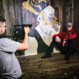 Mężczyzna filmujący siedzącego modela na tle drewnianej ściany z abstrakcyjnym rysunkiem