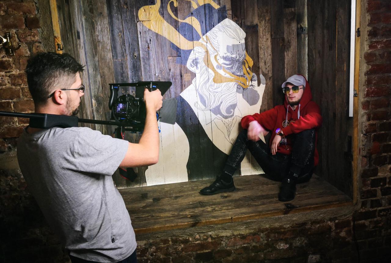 Mężczyzna filmujący siedzącego modela na tle drewnianej ściany z abstrakcyjnym rysunkiem