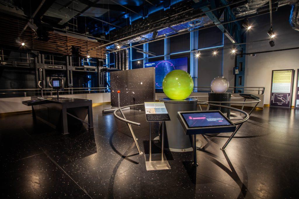 Wnętrze nowoczesnego centrum nauki z interaktywnymi eksponatami, w tym zielona kula na postumencie, ekrany dotykowe i panele informacyjne, oświetlone lampami punktowymi i niebieskim światłem z okien.