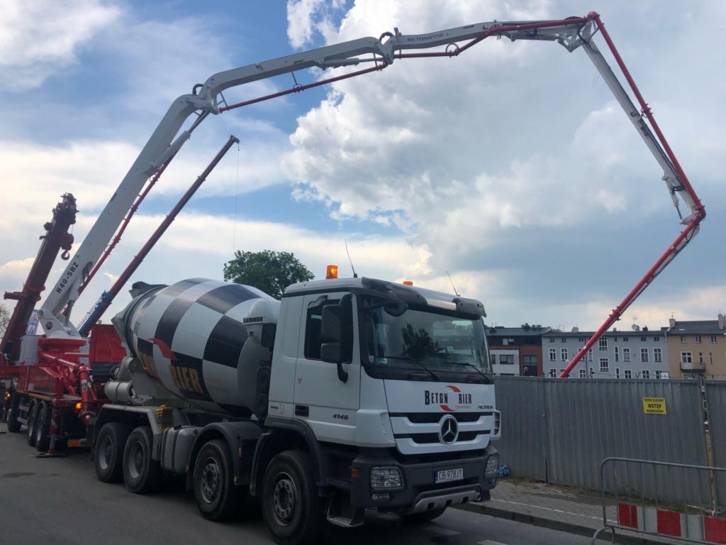 Dwa zestawy betonomieszarki z pompami do betonu na tle budowy i pochmurnego nieba. Na pierwszym planie ciężarówka z napisem 'BETON RIER' i bębnem w czarno-białą szachownicę.