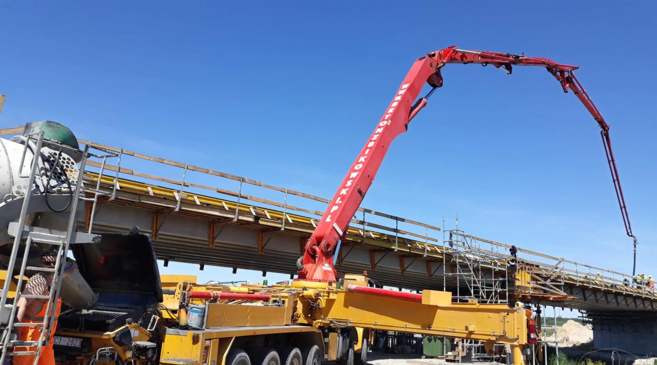 Żółty betonomieszarka z czerwoną pompą do betonu w trakcie wylewania betonu na konstrukcję mostu, widoczna drabina i pracownik w pomarańczowym kombinezonie.